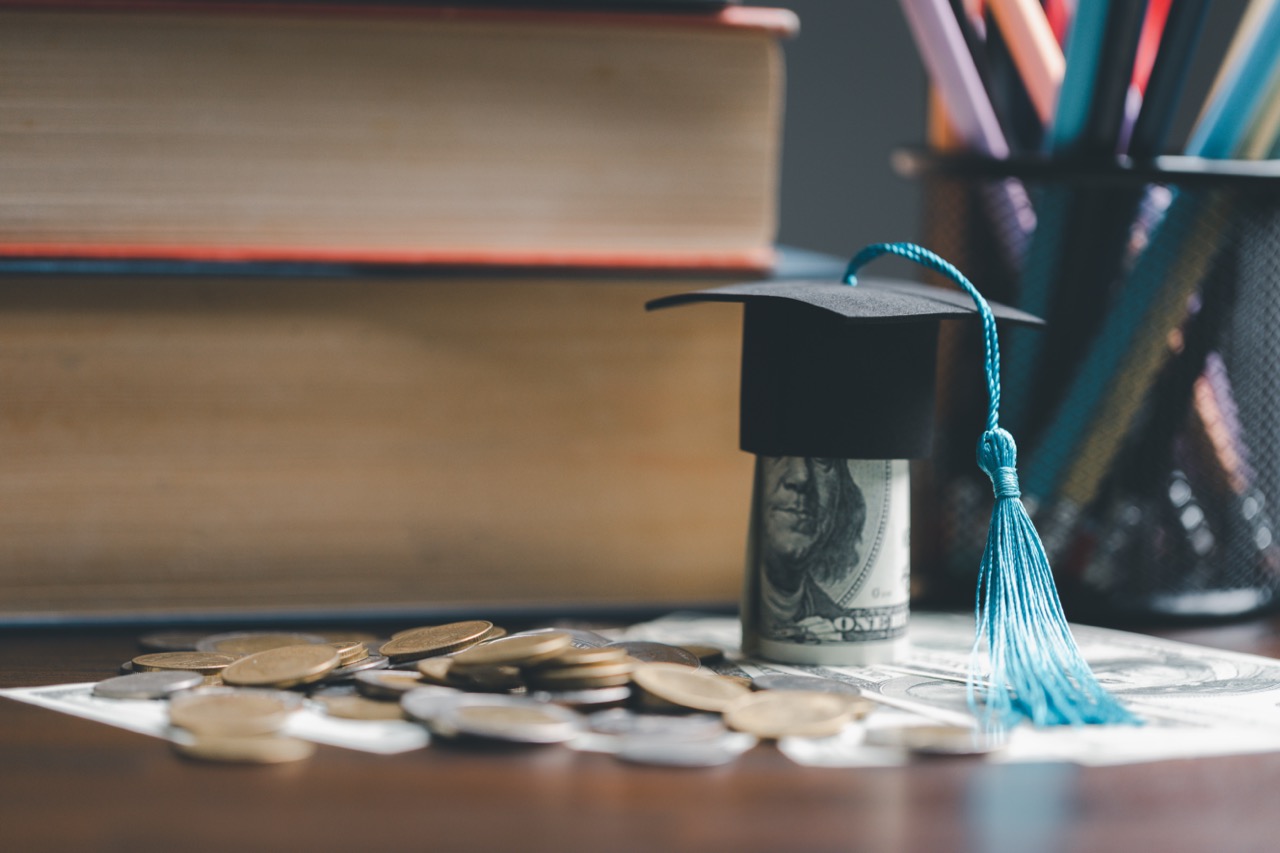 Graduation cap on a hundred dollar bill with coins and dollars blow it scattered around a table